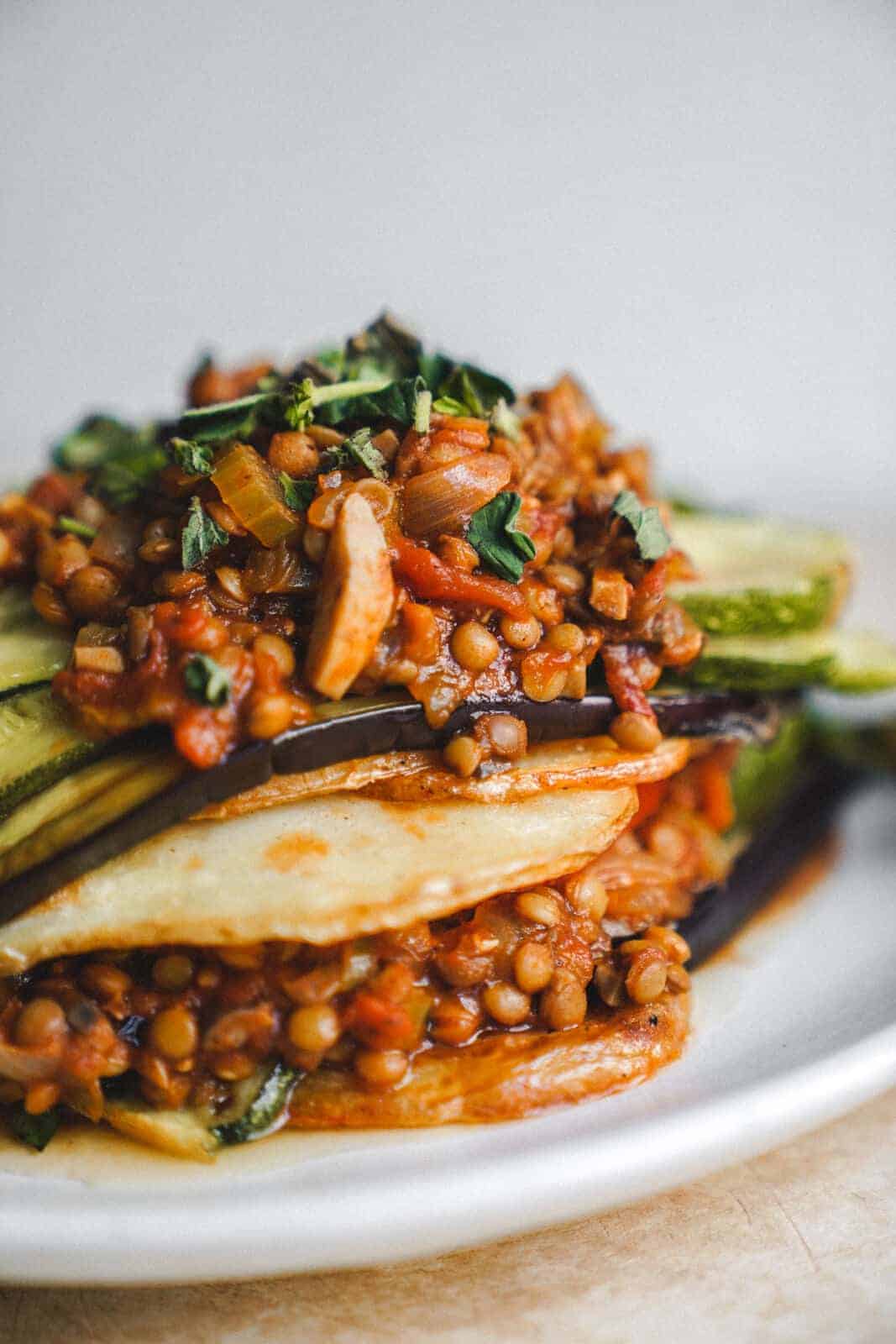 A close-up of a plate reveals a vegan moussaka: layers of zucchini and eggplant crowned with a savory lentil stew, diced tomatoes, and herbs. This colorful and hearty dish reimagines tradition in every delicious bite.