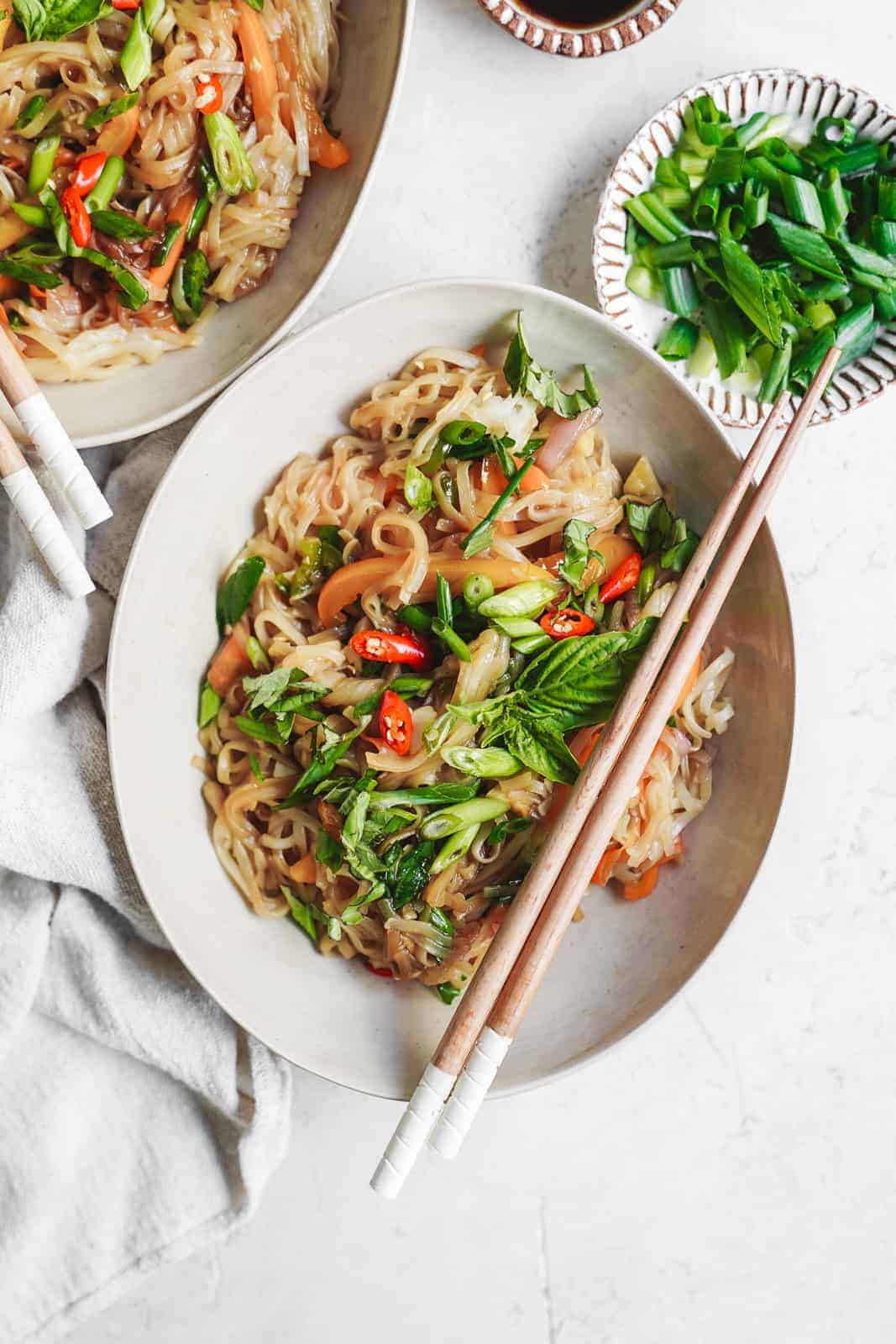 A bowl of Vegan Thai Drunken Noodles stir-fry with chopsticks rests on a light-colored table. It’s garnished with sliced red chilies, green onions, and fresh herbs. Nearby, a small bowl overflows with chopped green onions on a pristine white cloth.