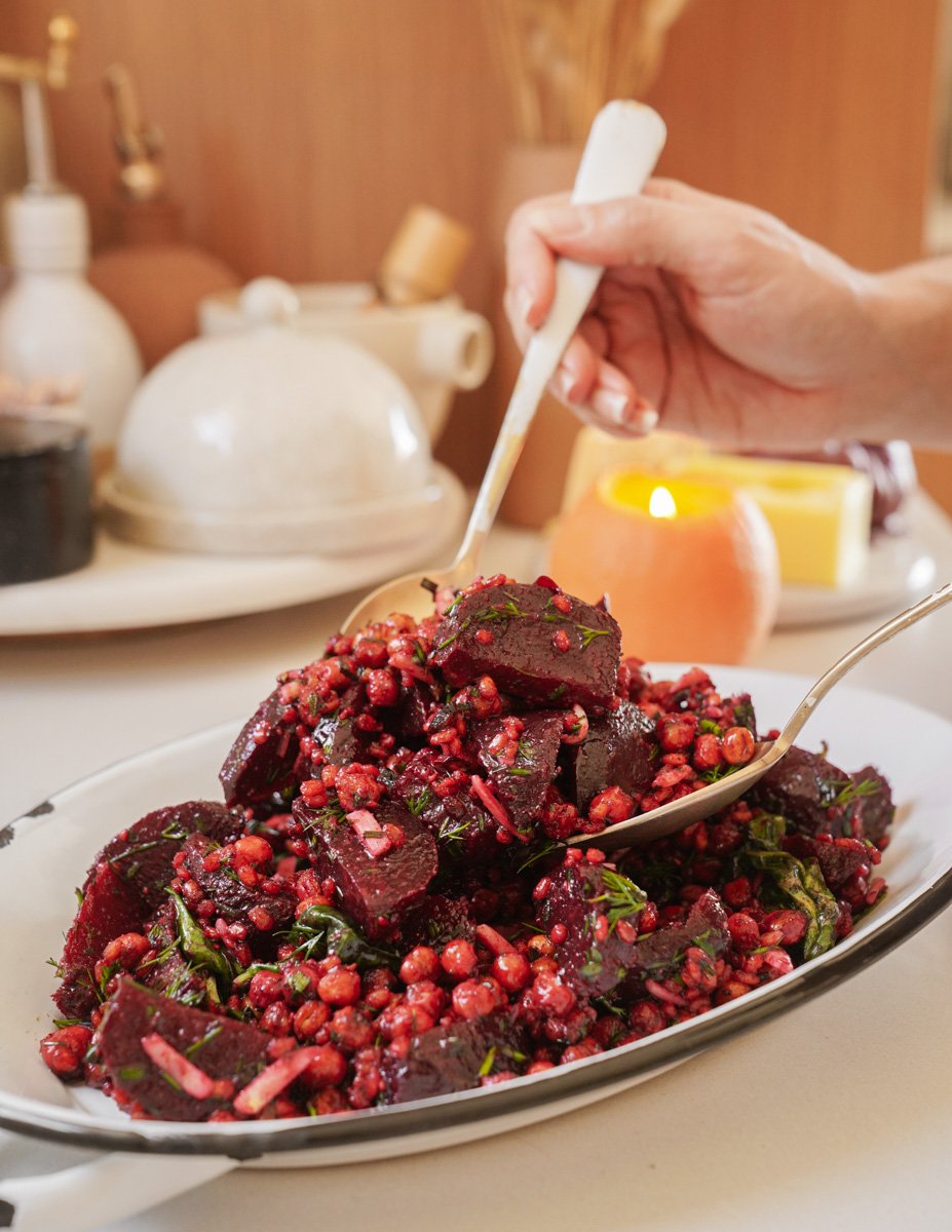 A hand serving a vibrant salad with roasted beets, pearl couscous, and greens on an oval platter. The background includes a teapot, candle, and condiments, creating a cozy dining atmosphere.