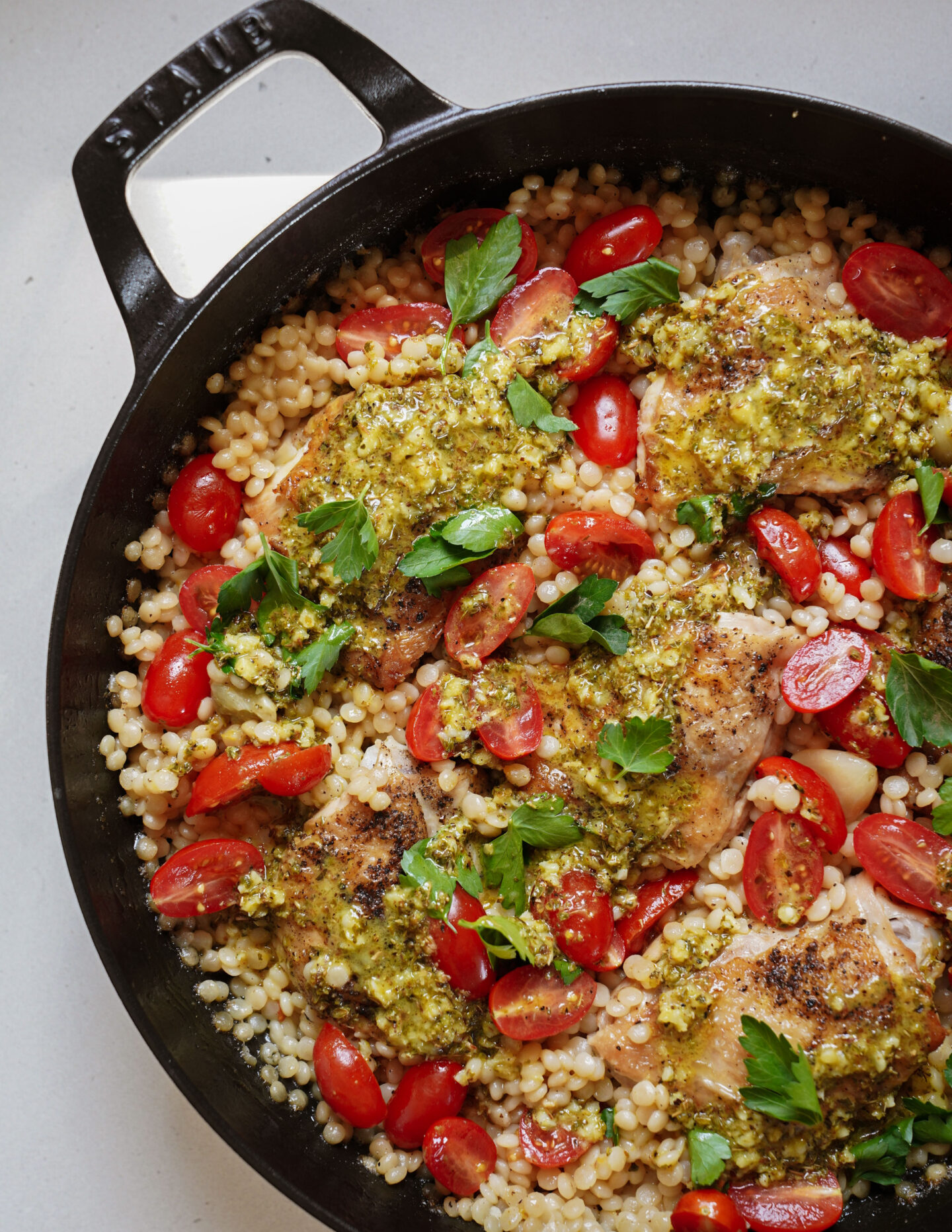 A skillet filled with cooked chicken thighs topped with a green pesto sauce, surrounded by couscous and halved cherry tomatoes. Garnished with fresh parsley leaves.