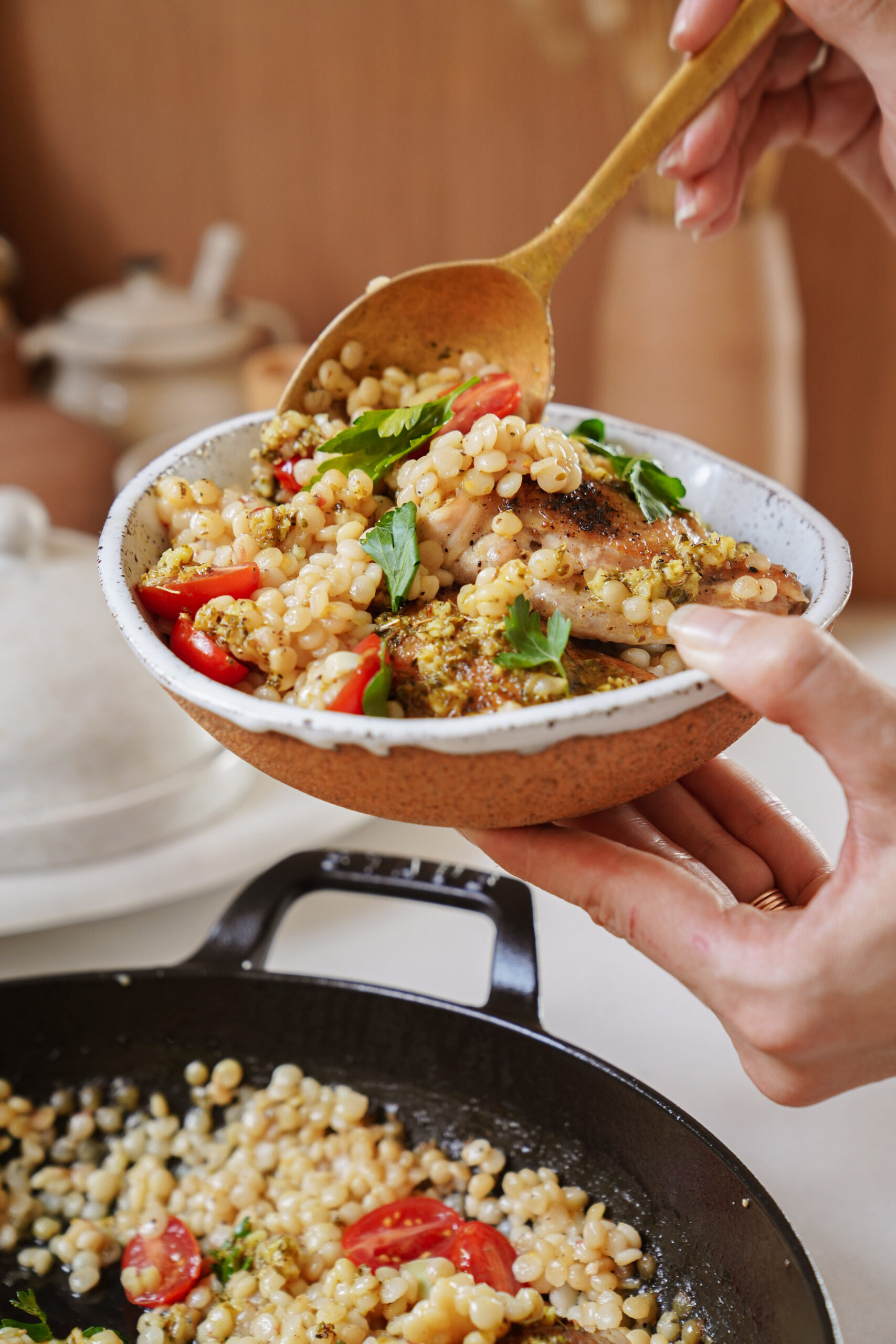 A person holding a bowl of couscous salad with cherry tomatoes and herbs, using a wooden spoon to serve it. A skillet with more salad is visible below. The setting is a kitchen countertop with neutral tones.