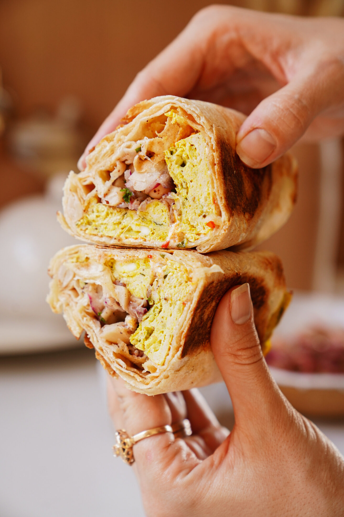 Close-up of hands holding a halved wrap sandwich filled with scrambled eggs, grilled chicken, and vegetables. The wrap shows a toasted exterior, with the vibrant filling visible. The background is softly blurred.