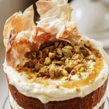 A close-up of a layered cake topped with whipped cream, a drizzle of honey, crumbled pistachios, and crispy phyllo pastry pieces, creating a textured and inviting appearance.