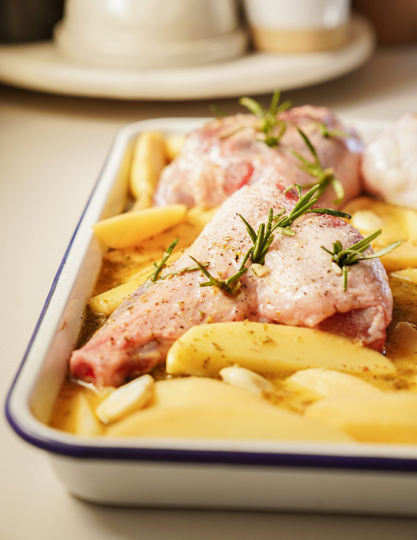 Raw lamb shank seasoned with herbs and pepper, laid on top of sliced potatoes and garlic cloves in a baking dish, ready to be cooked.