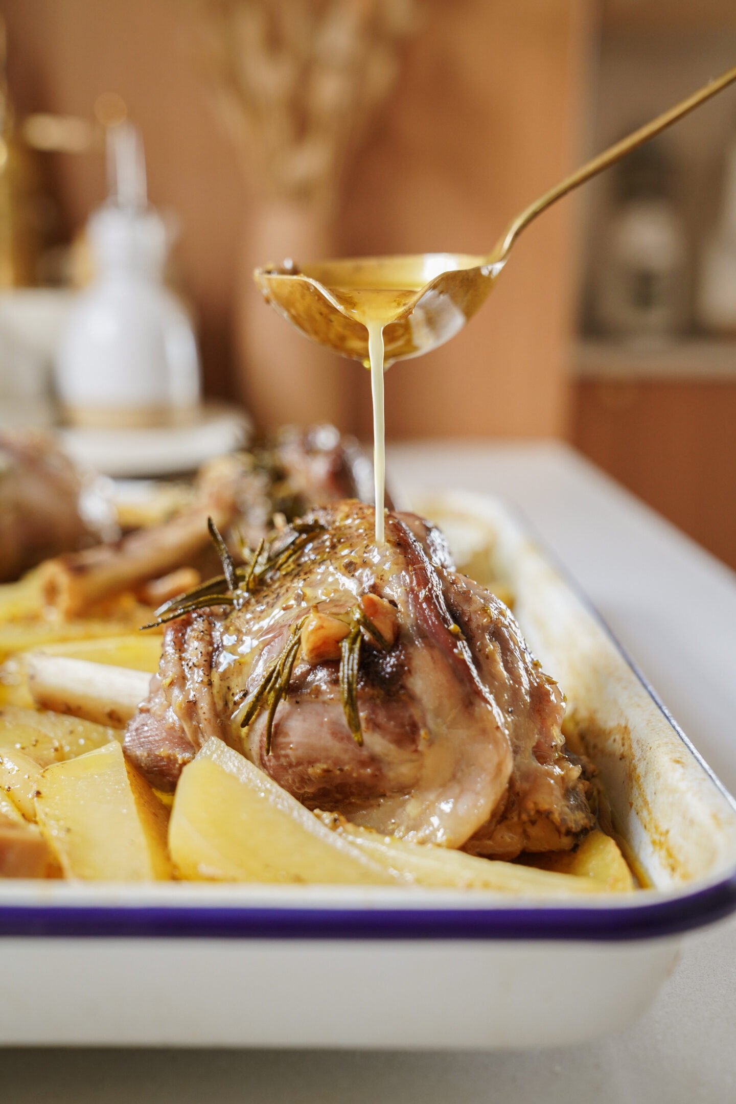 A close-up of a baked meat dish with rosemary and potatoes in a white tray, as a hand drizzles sauce over the meat with a spoon. The background is a softly focused kitchen scene.