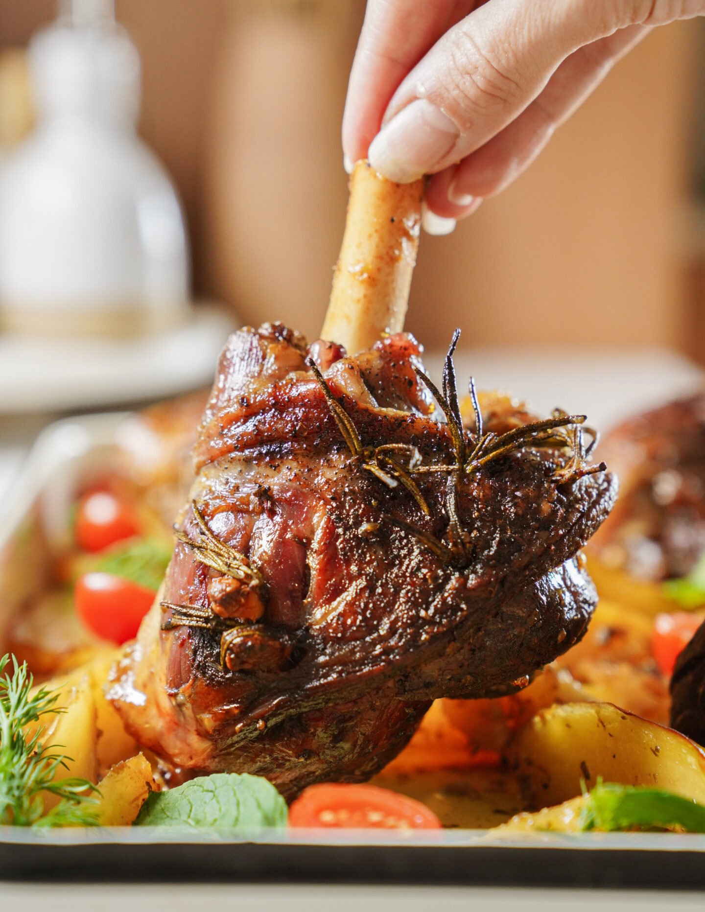 A hand holds a cooked lamb shank garnished with herbs, surrounded by roasted potatoes, cherry tomatoes, and fresh greens on a tray.