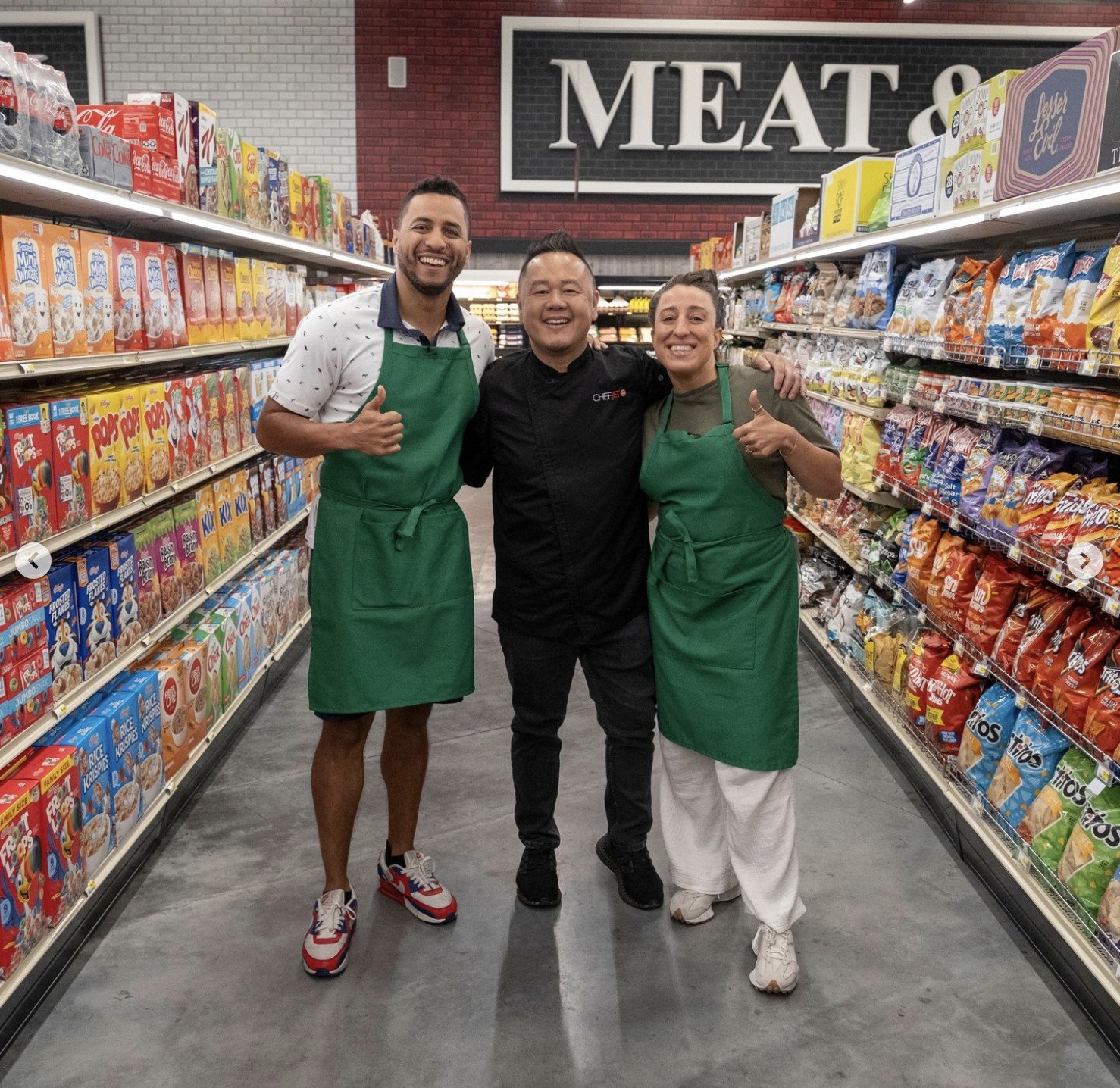 Three people stand happily in a grocery store aisle filled with cereal boxes and chips. Two wear green aprons and give thumbs up, while the middle person in black chef attire smiles. A large MEAT & sign is visible above.